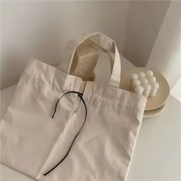 A Drawstring Canvas Shopping Bag in beige with a black string, customizable via OEM and ODM, is placed on a white table beside a plate holding a bubble candle.