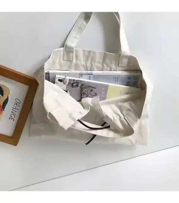 A cream-colored Drawstring Canvas Shopping Bag filled with magazines rests on a white surface beside a framed picture.