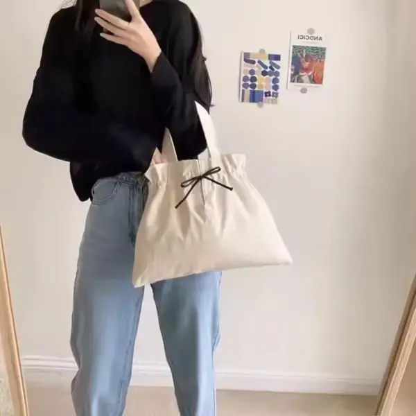 Person in a black top and light jeans holding a Drawstring Canvas Shopping Bag, featuring a white design with a black ribbon, taking a mirror selfie.
