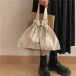 A person holding a Minimalist Canvas Drawstring Tote for Women in beige labeled "LOGO" e um saco de papel marrom, Vestindo uma saia preta e sapatos.