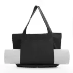 Women's Yoga Fitness Tote with shoulder straps, ಕಪ್ಪು ಬಣ್ಣದಲ್ಲಿ ತೋರಿಸಲಾಗಿದೆ, ಬಿಳಿ ಹಿನ್ನೆಲೆಯ ವಿರುದ್ಧ ಅದರ ಮುಂಭಾಗದ ಕಿಸೆಯಲ್ಲಿ ಸುತ್ತಿಕೊಂಡ ಯೋಗ ಚಾಪೆಯನ್ನು ಹೊಂದಿದೆ.